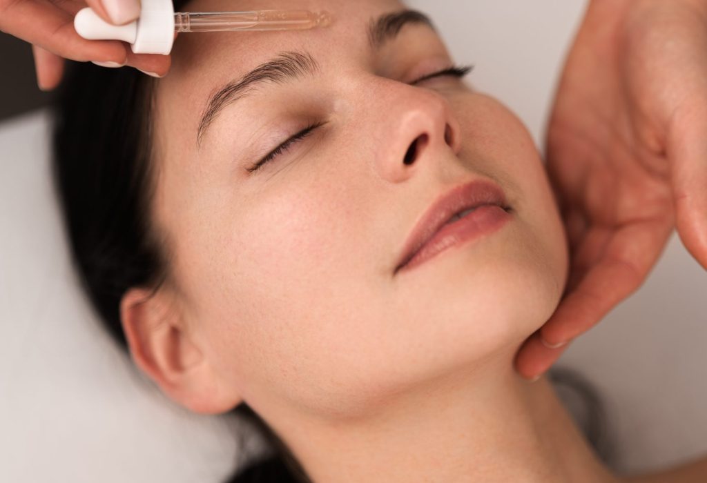 From above of crop unrecognizable beautician applying essential oil with pipette on face of relaxed young female client with dark hair, lying on table with closed eyes in beauty salon